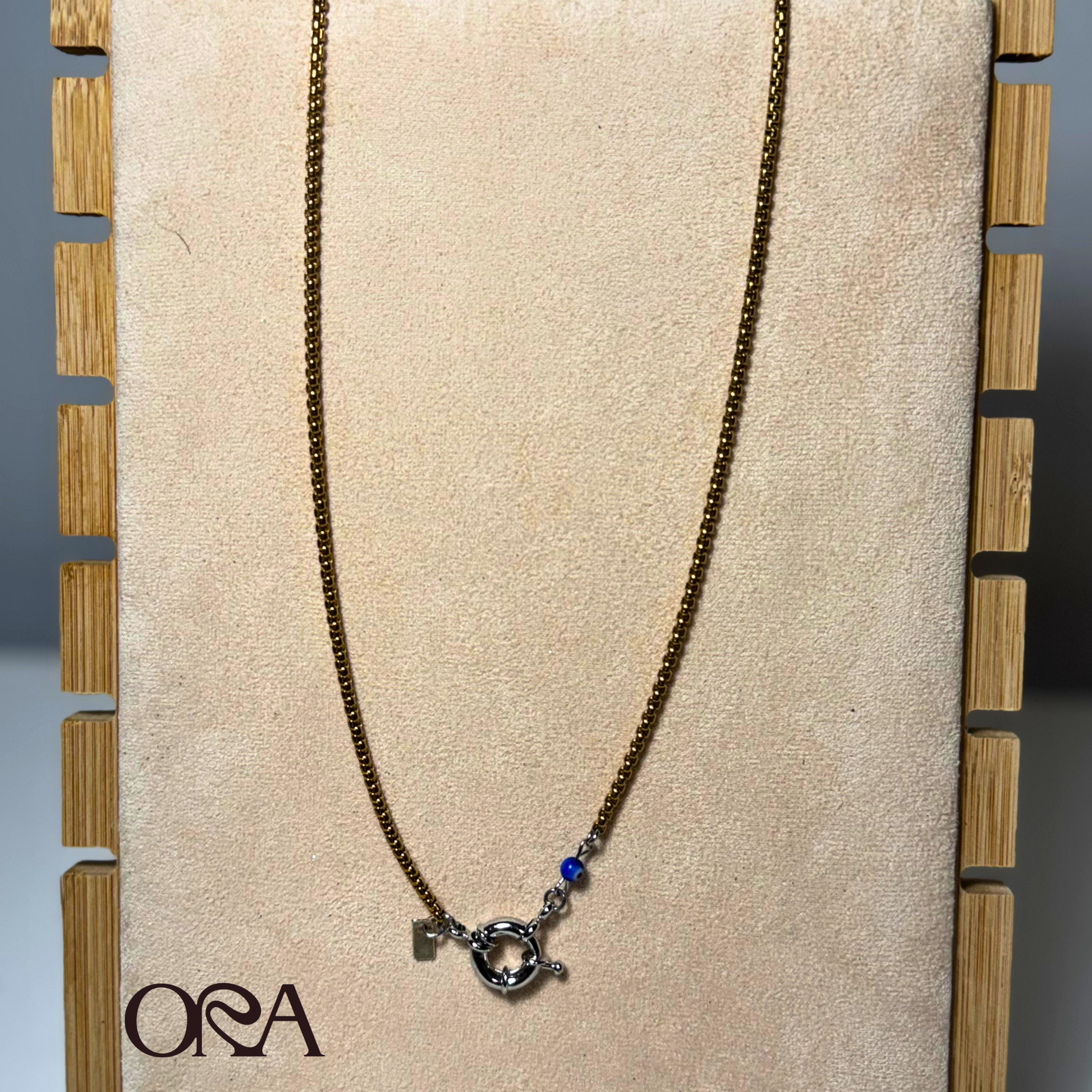 Box Chain Necklace in gold with silver claspon a beige board with 'ORA' branding