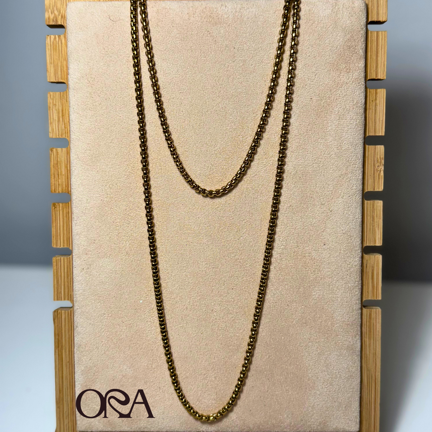 Gold chain necklace on a beige board with 'ORA' branding, displayed against a neutral background.