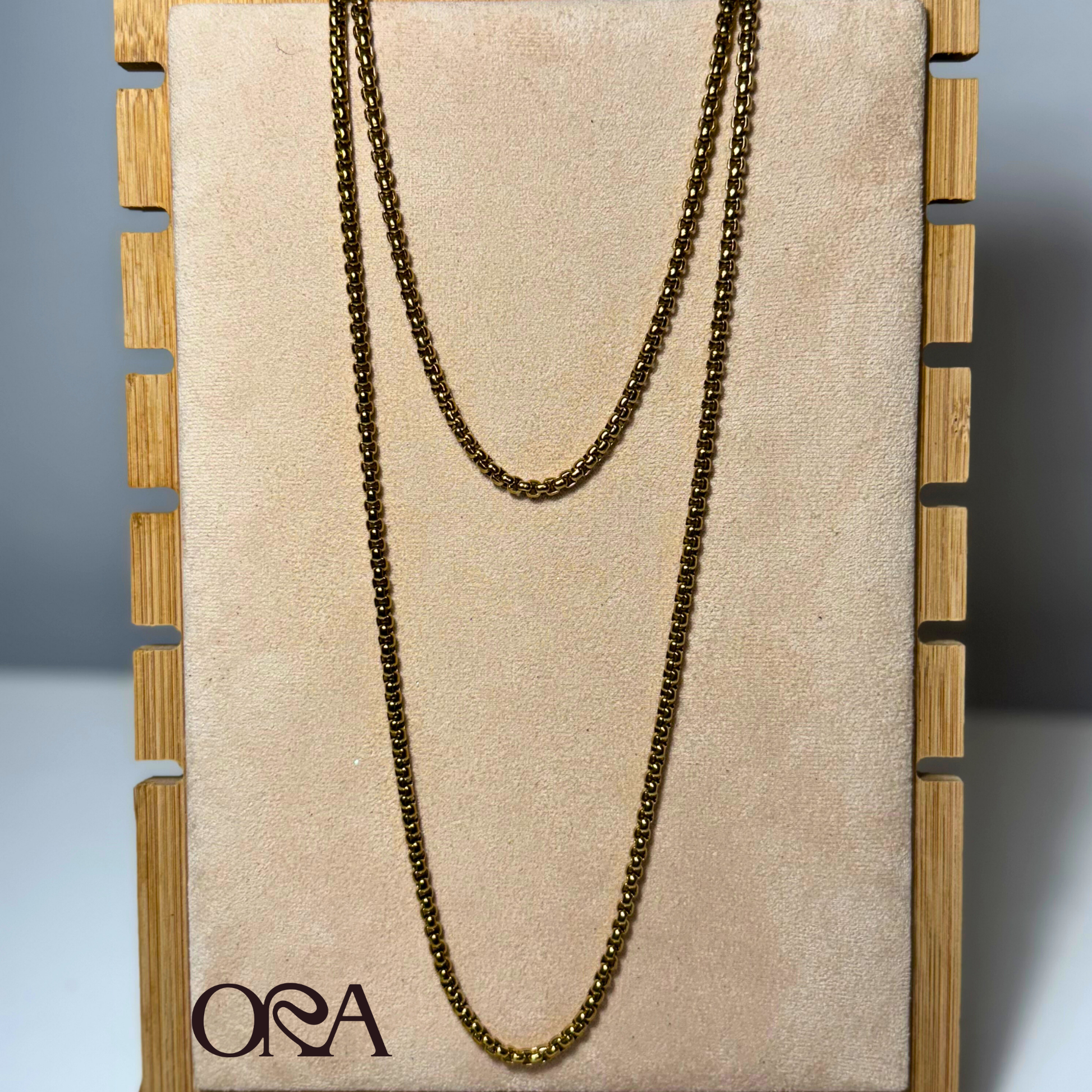 Gold chain necklace on a beige board with 'ORA' branding, displayed against a neutral background.