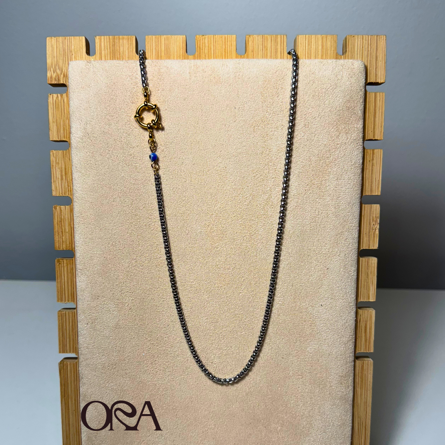 Necklace on a beige card with 'ORA' branding against a wooden stand.